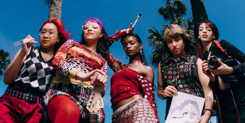 Five women posing together outdoors against a bright blue sky with palm trees in the background, wearing colorful patterned outfits and holding various objects.