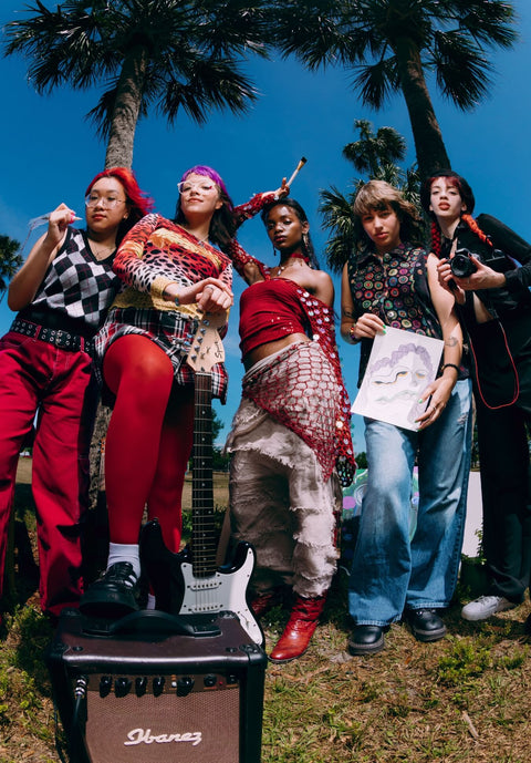 Five women posing outdoors with musical instruments under palm trees, including a black Ibanez amplifier in the foreground.